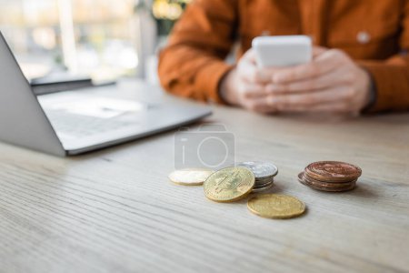 Photo for KYIV, UKRAINE - OCTOBER 18, 2022: selective focus of golden and silver bitcoins on desk near cropped businessman using mobile phone near laptop on blurred background in office - Royalty Free Image