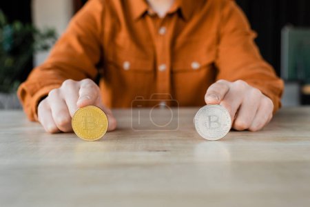 Photo for KYIV, UKRAINE - OCTOBER 18, 2022: partial view of accomplished businessman in trendy shirt sitting at office desk and touching golden and silver bitcoins, blurred background - Royalty Free Image