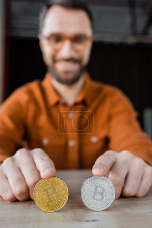 Foto de KYIV, UCRANIA - 18 DE OCTUBRE DE 2022: Barbudo exitoso hombre de negocios tocando bitcoins de plata y oro mientras está sentado en el escritorio en la oficina y sonriendo sobre un fondo borroso - Imagen libre de derechos