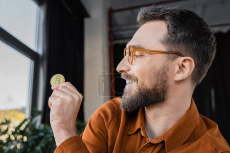 Photo for KYIV, UKRAINE - OCTOBER 18, 2022: brunette bearded businessman, pleased and successful, wearing trendy eyeglasses, looking at golden bitcoin in contemporary office on blurred background - Royalty Free Image