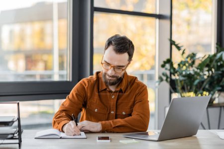 positive bearded businessman in eyeglasses and trendy shirt writing in notebook near laptop, smartphone with blank screen, documents tray and sticky notes at workplace in office
