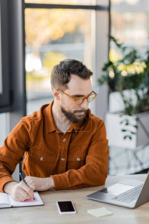 konzentrierter Geschäftsmann mit trendiger Brille und Hemd, der in ein Notizbuch neben Laptop und Handy mit leerem Bildschirm am Arbeitsplatz im modernen Büro schreibt