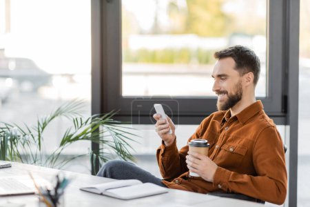 happy and successful bearded businessman with takeaway drink messaging on mobile phone near notebook, laptop, decorative plants and large windows in office