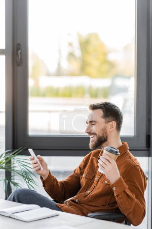 überglücklicher Geschäftsmann im trendigen Hemd, Kaffee in Pappbecher haltend und beim Sitzen am Arbeitsplatz neben großen Fenstern im modernen Büro auf das Handy blickend