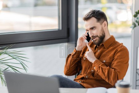 hombre de negocios barbudo positivo y carismático con camisa elegante que habla en el teléfono móvil mientras está sentado cerca de la computadora portátil borrosa y ventanas en la oficina contemporánea