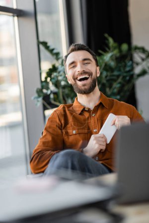 aufgeregter und erfolgreicher Unternehmer im trendigen Hemd, Handy in der Hand und lachend in die Kamera neben verschwommenem Laptop am Arbeitsplatz im modernen Büro