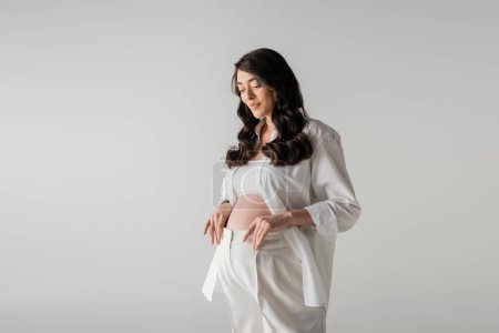 Charmante und glückliche schwangere Frau mit welligen brünetten Haaren posiert in weißem Hemd, bauchfreiem Oberteil und Hose, während sie isoliert auf grauem Hintergrund lächelt, Umstandsmodekonzept