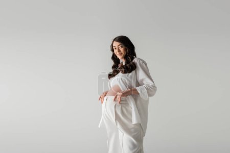 glücklich zukünftige Mutter mit welligen brünetten Haaren posiert in weißen modischen Kleidern wie weißem Hemd und Hose isoliert auf grauem Hintergrund, trendiges Umstandskonzept
