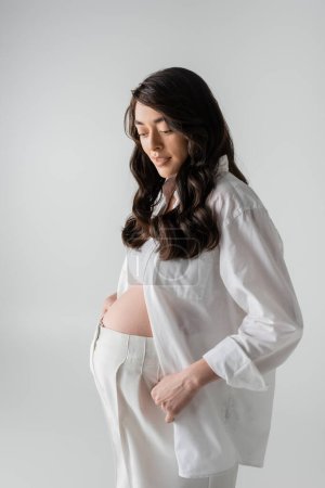 charming future mother in white fashionable clothes such as white shirt, crop top and pants smiling isolated on grey background, stylish maternity concept, pregnancy 