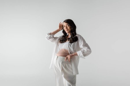 fashionable pregnant woman with wavy brunette hair posing in white pants and shirt while smiling at camera isolated on grey background, maternity style concept