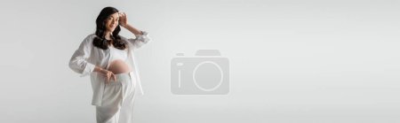 hübsche und glückliche Schwangere in weißem Hemd, bauchfreiem Oberteil und Hose, die welliges brünettes Haar fixiert und isoliert auf grauem Hintergrund in die Kamera blickt, trendiges Umstandskonzept, Banner