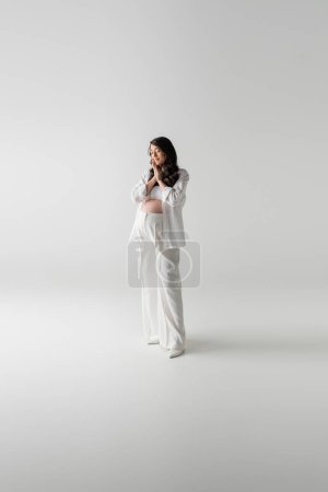 full length of trendy future mother in white fashionable pants and shirt standing with closed eyes and smiling on grey background, maternity style concept, pregnancy 