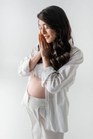 charming and pleased pregnant woman with wavy brunette hair, wearing white crop top, shirt and pants, holding hands near face and smiling isolated on grey background, trendy maternity concept