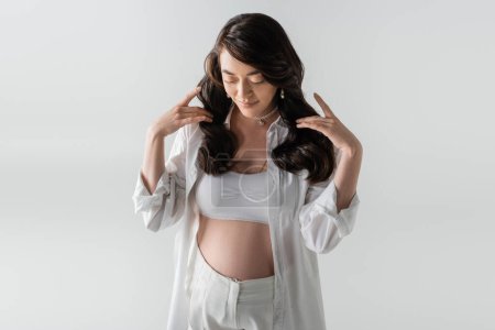 fröhliche Schwangere in weißem Crop Top, stylischem Hemd und Hose, die welliges brünettes Haar berührt und isoliert auf grauem Hintergrund nach unten schaut, Umstandsmodekonzept