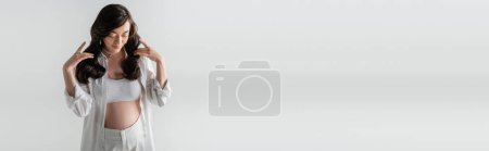stylische Schwangere in weißem, trendigem Hemd, bauchfreiem Oberteil und Hose, die welliges brünettes Haar berührt und isoliert auf grauem Hintergrund posiert, modisches Umstandskonzept, Banner