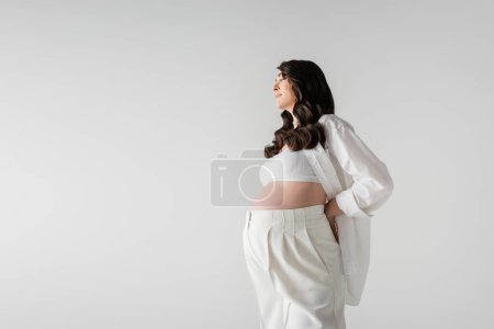 Téléchargez les photos : Vue latérale de la femme enceinte souriante aux cheveux bruns ondulés, portant des vêtements de maternité blancs élégants, posant avec la main derrière le dos isolé sur fond gris, concept de maternité à la mode - en image libre de droit