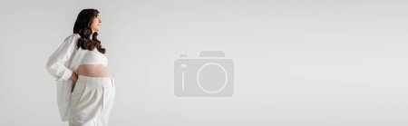 Foto de Vista lateral de modelo embarazada con cabello castaño ondulado posando en camisa elegante blanca y pantalones aislados sobre fondo gris, concepto de moda de maternidad, pancarta, moda de maternidad - Imagen libre de derechos