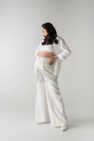 volle Länge der ansprechenden und glücklichen zukünftigen Mutter in weißen trendigen Hosen, bauchfreiem Oberteil und Hemd, das den Bauch auf grauem Hintergrund berührt, modische Mutterschaft, Schwangerschaft 