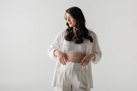 pretty expecting mother in white pants, crop top and stylish shirt holding hands near tummy, smiling and looking away isolated on grey background, maternity fashion concept