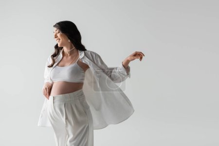 positive brunette pregnant woman in shirt, crop top and pants posing with outstretched hand isolated on grey background, fashionable pregnancy concept