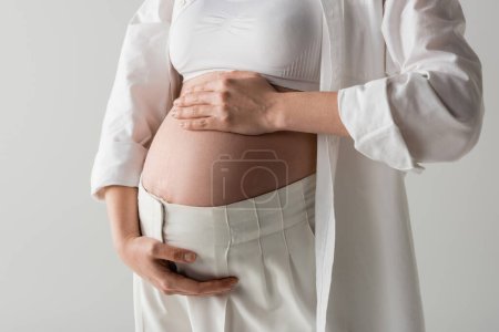 Teilansicht der zukünftigen Mutter in weißem, stylischem Hemd, bauchfreiem Oberteil und Hose, die den Bauch zärtlich umarmt, isoliert auf grauem Hintergrund, Umstandsmodekonzept, schwangere Frau 