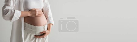 partial view of future mother in white crop top, shirt and pants embracing belly while standing isolated on grey background, maternity fashion concept, banner, pregnant woman 