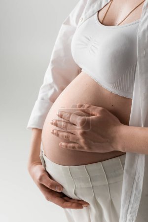 Ausgeschnittene Ansicht einer stilvollen werdenden Mutter in weißem bauchfreiem Top, Hemd und Hose umarmen den Bauch, während sie isoliert auf grauem Hintergrund steht, Umstandsmodekonzept, schwangere Frau 