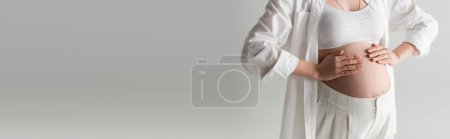 cropped view of trendy pregnant woman touching belly while posing in white crop top, shirt and pants isolated on grey background, maternity fashion concept, banner 
