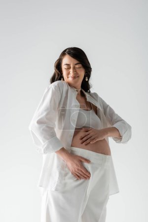 overjoyed future mother with closed eyes and pleased face expression, wearing white stylish shirt and pants, touching tummy isolated on grey background, maternity fashion concept