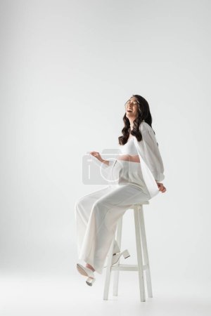 full length of excited pregnant woman with wavy brunette hair sitting on stool in white trendy clothes and smiling with closed eyes on grey background, maternity fashion concept
