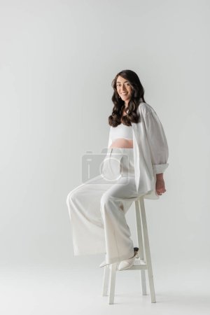 full length of pleased expecting mother in white fashionable pants, crop top and shirt sitting on stool and looking at camera on grey background, maternity fashion concept, pregnant woman 