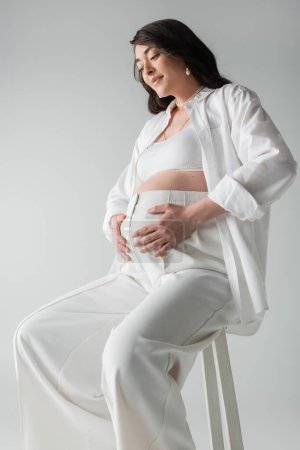 lächelnde und ansprechende Schwangere in weißem Hemd, bauchfreiem Oberteil und Hose auf Hocker sitzend und den Bauch isoliert auf grauem Hintergrund umarmend, Umstandskonzept, werdende Mutter
