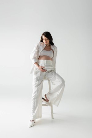full length of happy mom-to-be in white pants, crop top and shirt posing near stool and touching tummy on grey background, fashionable pregnancy concept, pregnant woman 