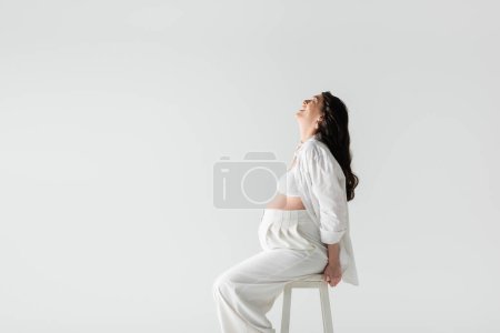 Téléchargez les photos : Vue latérale de joyeuse femme enceinte avec des cheveux bruns ondulés assis sur des tabourets en chemise blanche, haut de culture et pantalon isolés sur fond gris, concept de maternité tendance - en image libre de droit