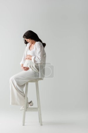 volle Länge der stilvollen zukünftigen Mutter in weißem Hemd und weißer Hose auf Hocker sitzend und zärtlich umarmend Bauch auf grauem Hintergrund, Umstandsmodekonzept