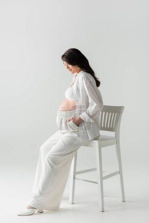 volle Länge der ansprechenden Schwangeren in bauchfreiem Top und Hose auf Stuhl sitzend mit der Hand in Hosentasche auf grauem Hintergrund, Umstandskonzept