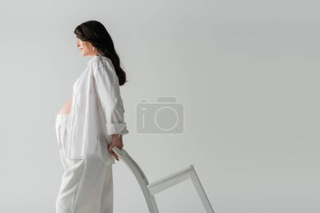 Foto de Vista lateral de la mujer embarazada de moda en camisa blanca y pantalones posando con silla mientras está de pie aislado sobre fondo gris, concepto de moda de maternidad - Imagen libre de derechos