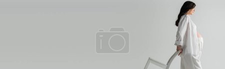 Foto de Vista lateral de la sonriente futura madre con cabello castaño ondulado, con camisa blanca y pantalones, posando con silla sobre fondo gris, elegante concepto de maternidad, pancarta - Imagen libre de derechos