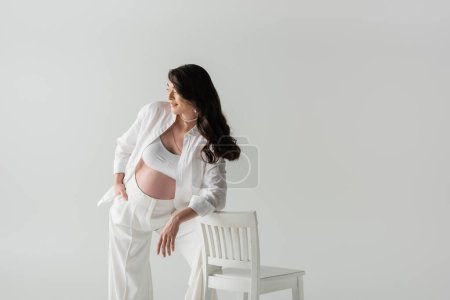 zufriedene Schwangere in modischen Hosen, bauchfreiem Oberteil und Hemd auf Stuhl gestützt und vereinzelt auf grauem Hintergrund wegschauend, Umstandsmodekonzept