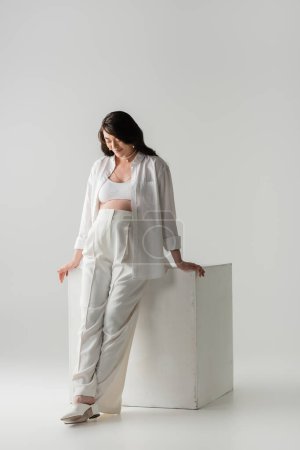 full length of happy pregnant woman with wavy brunette hair, wearing pants, shirt and crop top, smiling near white cube on grey background, maternity fashion concept