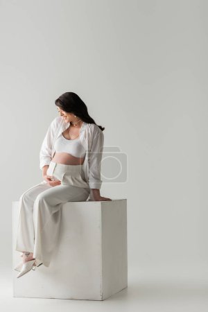 volle Länge der lächelnden werdenden Mutter in stylischer Hose, bauchfreiem Oberteil und Hemd, die den Bauch berührt, während sie auf einem weißen Würfel auf grauem Hintergrund im Studio sitzt, Umstandsmodekonzept, schwangere Frau 