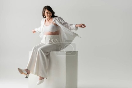 full length of cheerful pregnant woman in fashionable pants, shirt and crop top sitting on white cube on grey background, maternity fashion concept