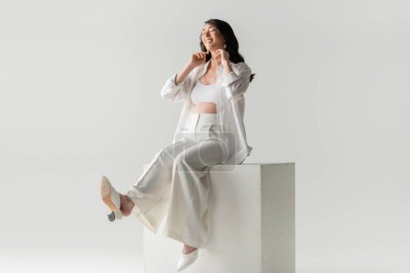 full length of joyful expecting mother in trendy pants, shirt and cropped top sitting on white cube and smiling with closed eyes on grey background, maternity fashion concept