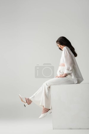 Téléchargez les photos : Vue latérale de la belle-mère brune heureuse en vêtements à la mode tels que chemise blanche et pantalon assis sur cube en studio sur fond gris, concept de mode maternité - en image libre de droit