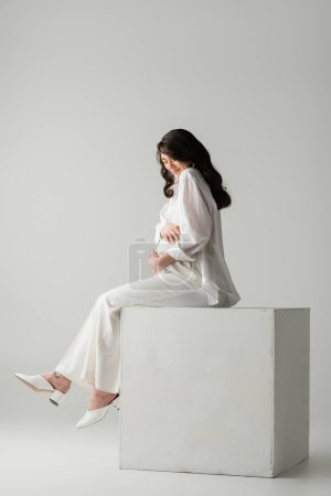 Téléchargez les photos : Pleine longueur de femme enceinte brune en chemise blanche et pantalon embrassant ventre tout en étant assis sur cube en studio sur fond gris, concept de mode maternité - en image libre de droit