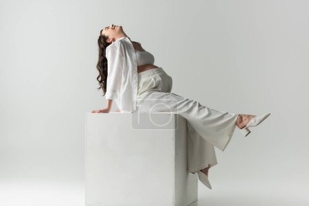 Full length of excited future mother in white stylish clothes such as pants, crop top and shirt sitting on white cube on grey background, maternity fashion concept