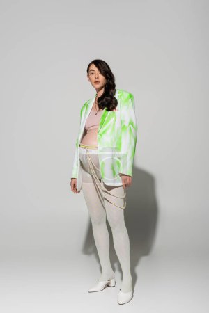 longueur totale de la future mère en leggings, crop top, blazer vert et blanc et ceinture de perles debout et regardant la caméra sur fond gris, concept de grossesse élégant