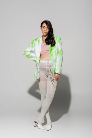 full length of pretty brunette mother-to-be in green and white jacket, crop top, beads belt and tights posing with hand on hip and looking at camera on grey background, trendy pregnancy concept