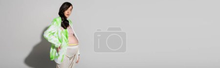 Foto de Elegante mamá-a-ser con el pelo ondulado morena, usando chaqueta verde y blanco, leggins y abalorios cinturón de pie con la mano en la cadera sobre fondo gris, concepto de moda de maternidad, pancarta, mujer embarazada - Imagen libre de derechos