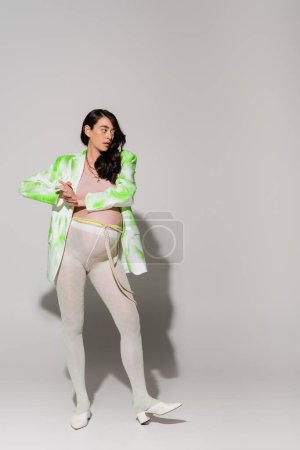 in voller Länge stilvolle werdende Mutter in grün-weißer Jacke, bauchfreiem Oberteil, Leggings und Perlengürtel auf grauem Hintergrund, modisches Schwangerschaftskonzept, schwangere Frau 